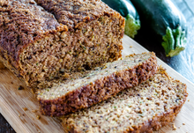 Load image into Gallery viewer, A loaf of fresh-baked zucchini bread sits on a wooden cutting board. Two slices are laying in front of the rest of the loaf. Two fresh zucchinis are to right right of the cutting board.
